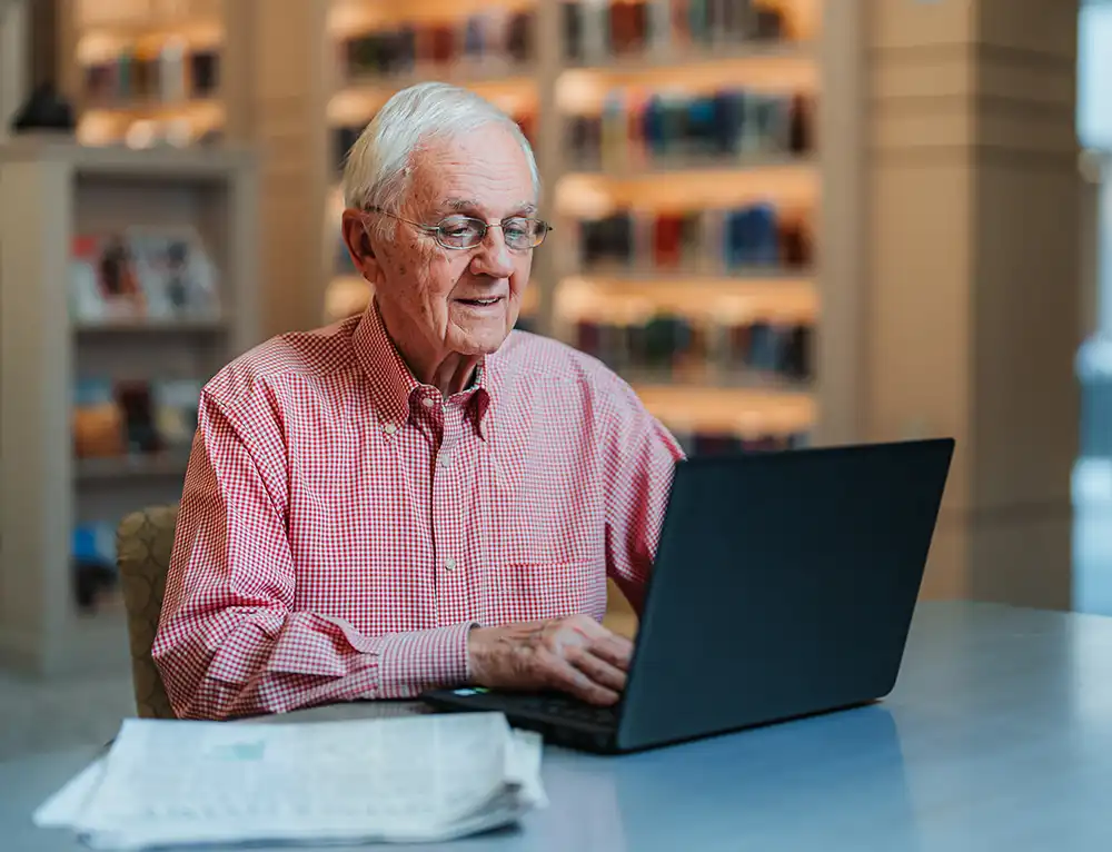 A senior man sits at a table in a library using a laptop