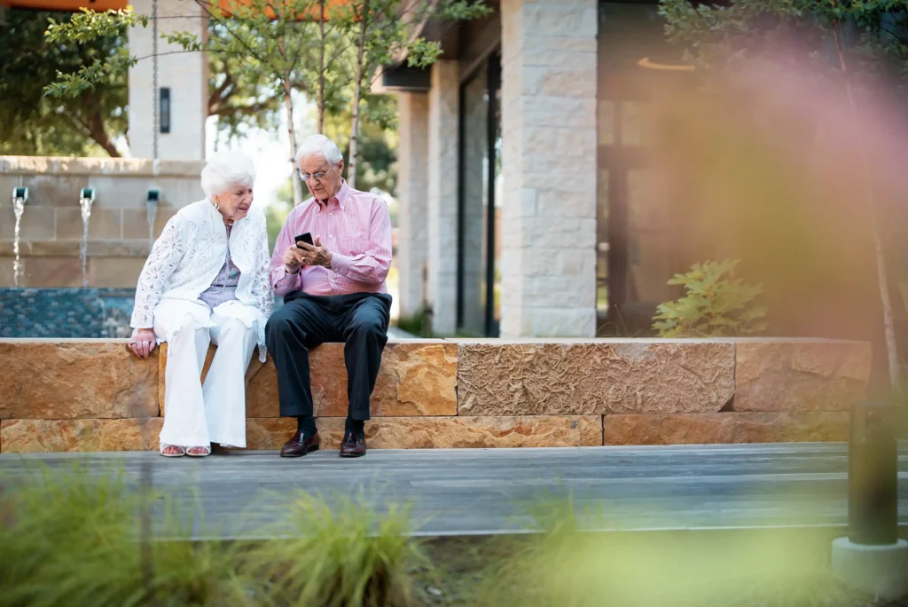 Two senior adults is on a wall by a water feature looking at a smartphone