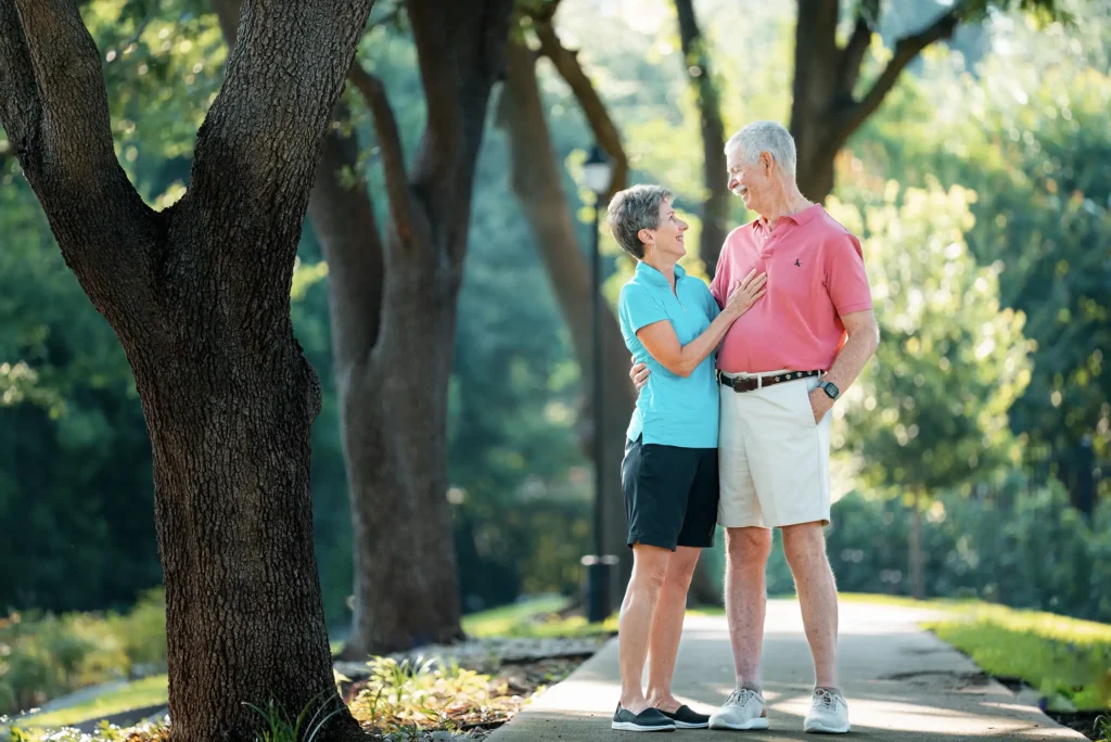 A senior couple stands together on an outdoor walkway surrounded by trees