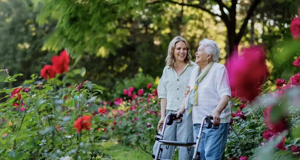 Retired mother and adult daughter walking outside among some rose bushes.